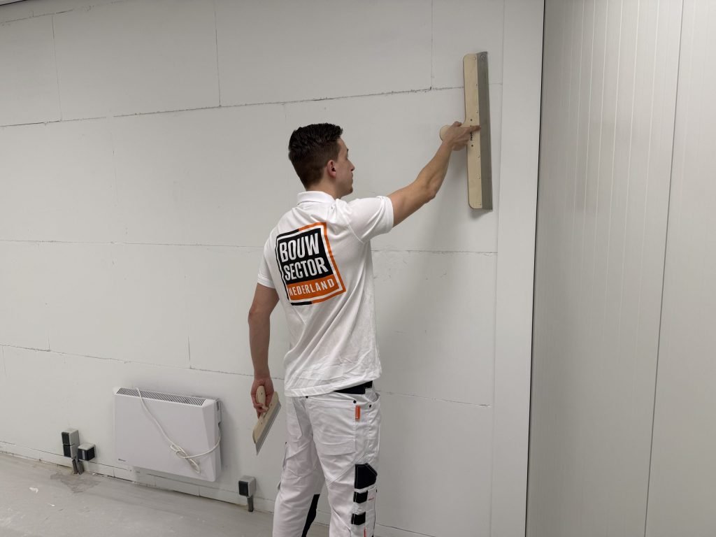 Vakman in wit werkshirt zet een brede spaan hoog tegen een wand van platen, plamuurmes in de andere hand