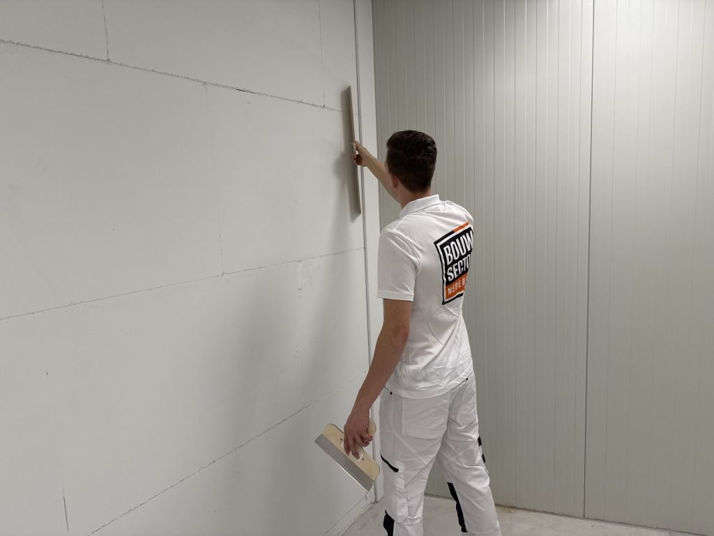 Jonge stukadoor houdt een spaan tegen een wand met zichtbare plaatnaden, met een plamuurmes in zijn linkerhand