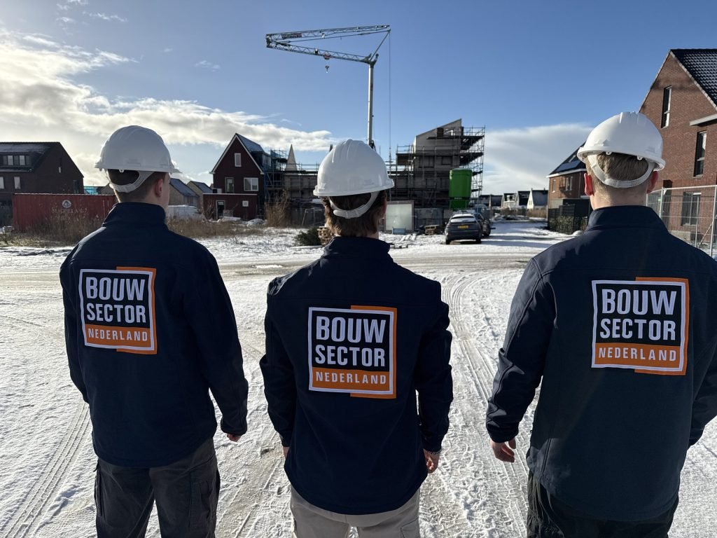 Drie vakmensen van Bouwsector Nederland met witte helmen kijken uit over een bouwplaats met nieuwe woningen in de sneeuw