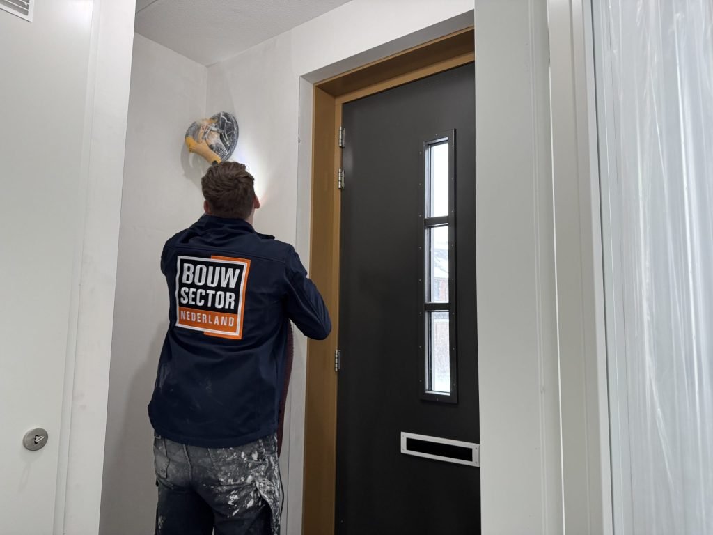Vakman van Bouwsector Nederland met een wandschuurmachine bij een voordeur met zijlicht