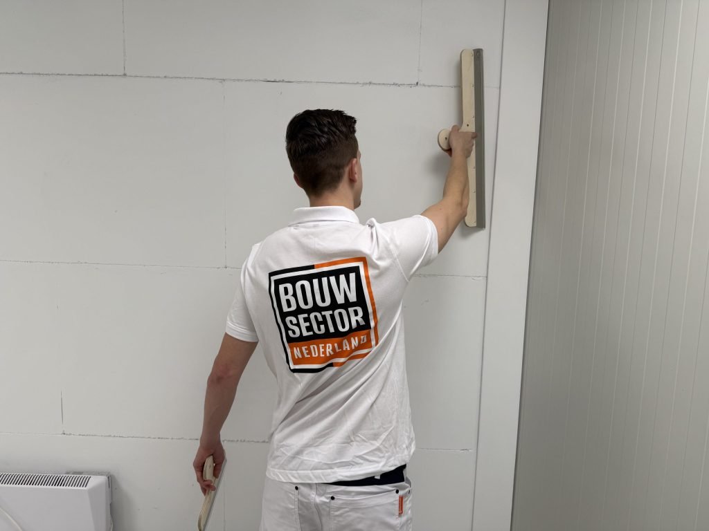 Stukadoor van Bouwsector Nederland houdt een lange spaan tegen de bovenkant van een witte wand
