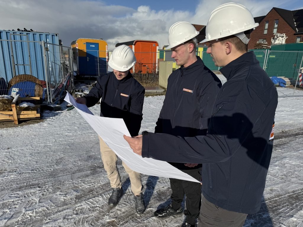 Drie vakmensen van Bouwsector Nederland met witte helmen kijken samen naar een bouwtekening op een bouwterrein